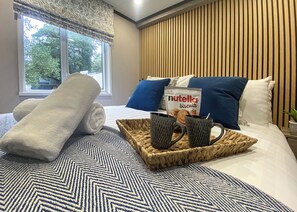 Un lit bien fait, recouvert d’une courtepointe à motifs, une serviette blanche roulée, un plateau avec un pot de Nutella et deux tasses noires.
