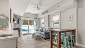 A spacious living area with a wooden floor, a gray sofa, and a dining table with stools. - Shiner Shore Shack: 1200 Ft to Beach Access, Pool, Hot Tub (Corolla)