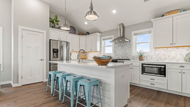 A modern kitchen with white cabinets, a stainless steel refrigerator, a microwave, and a large island with stools. - Shiner Shore Shack: 1200 Ft to Beach Access, Pool, Hot Tub (Corolla)