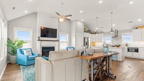 A spacious living room with a fireplace, a blue armchair, and a wooden dining table. - Shiner Shore Shack: 1200 Ft to Beach Access, Pool, Hot Tub (Corolla)
