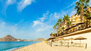 Un hotel frente a la playa con tumbonas y sombrillas, piscina y vistas al mar y a las montañas.