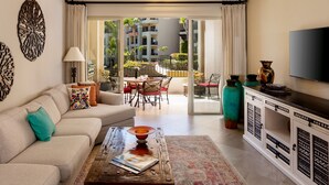 Una sala de estar moderna con un sofá, una mesa de centro y una televisión. Hay un balcón con vistas a edificios y zonas verdes.