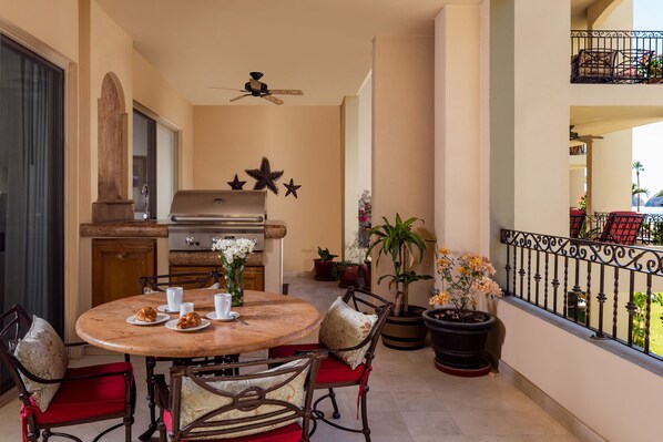 A dining area with a round table, chairs, and a grill. There are potted plants and a balcony with a railing.