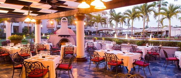 An outdoor dining area with tables and chairs, a central fire feature, and palm trees in the background. - Luxury Cabo San Lucas Vacation Rental at Villa La Estancia | Beachfront Condo, All-Inclusive Option (Cabo San Lucas)