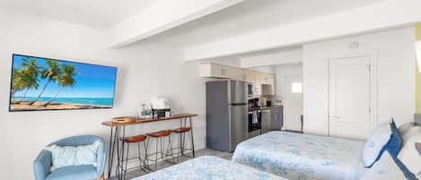 A hotel room with two beds, a desk with a chair, a kitchen area, and a painting of a beach.