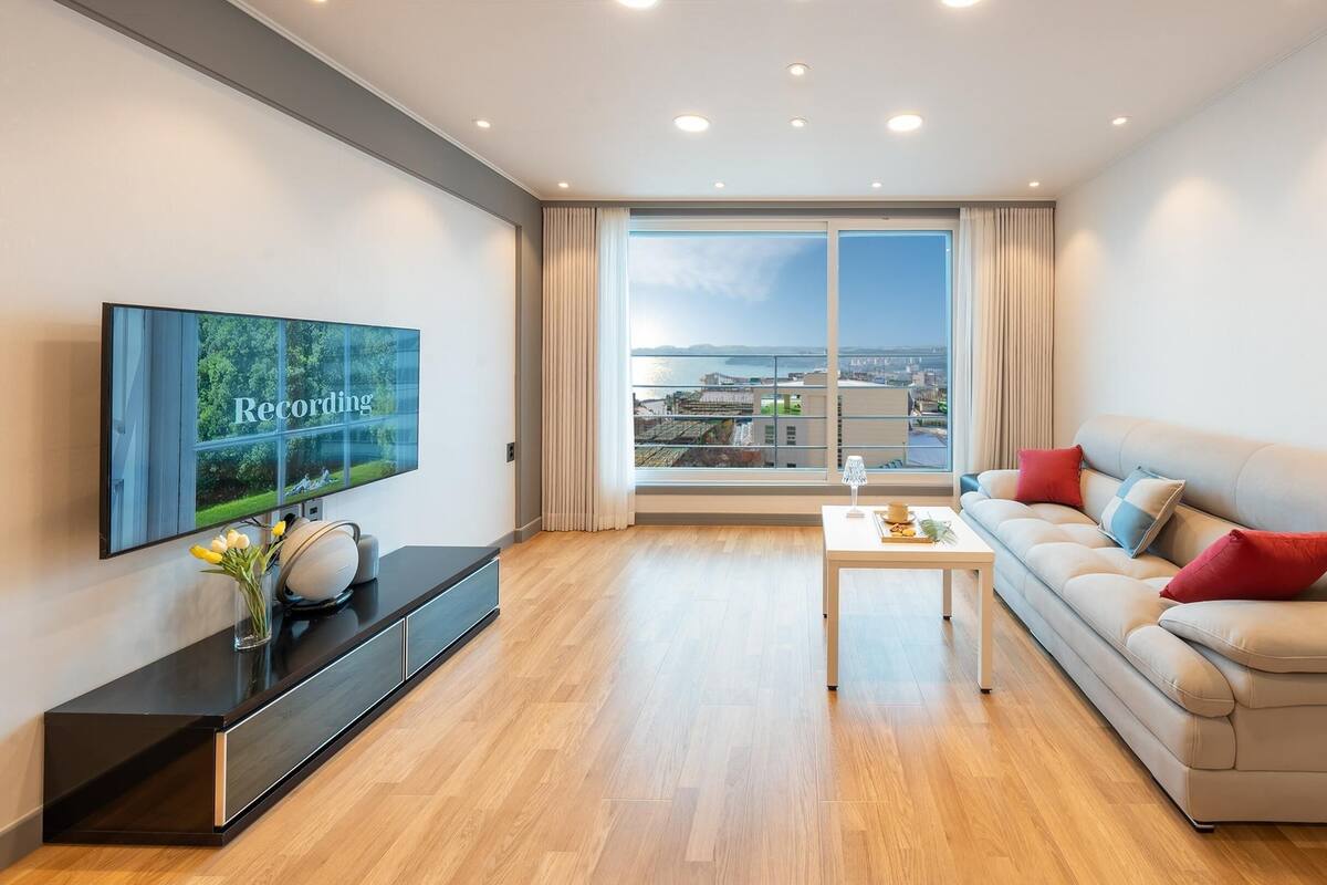 A modern living room with a large TV, a sofa, a coffee table, and a view of a cityscape through large windows.
