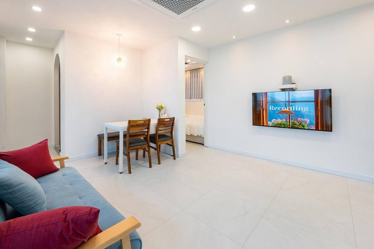 A modern living room with a sofa, a dining table with chairs, and a flat-screen TV mounted on the wall.