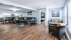Un vaste espace de vie comprenant un canapé de couleur sombre, une table basse et un fauteuil en osier. On aperçoit à l’arrière-plan un coin repas avec une table et des chaises.