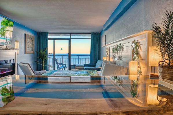 A modern hotel room with a glass coffee table, a bed with a blue and white striped bedspread, a sofa, a TV, and a view of the ocean.