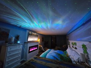 A room with a starry ceiling projection, a bed with a tropical-themed bedspread, a fireplace, and a television displaying a sports event.