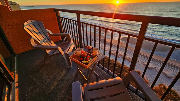 A balcony with two Adirondack chairs, a small table with fruits and a mug, and a view of the sunset over the ocean. - Oceanfront King Beach Suite- Updated Landmark 1030 (Myrtle Beach)