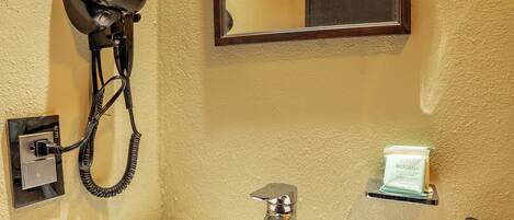 A bathroom with a white sink, a hairdryer mounted on the wall, a mirror, and a small shelf with a bottle.