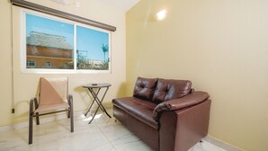 A room with a brown leather sofa, a beige armchair, a small round table, and a window with a view of a building and palm trees. - Hotel Guitarra Inn (Puerto Escondido)