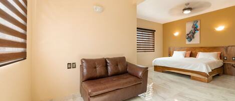 Una habitación de hotel moderna con un sofá de cuero marrón, una cama grande con ropa blanca y un ventilador de techo.