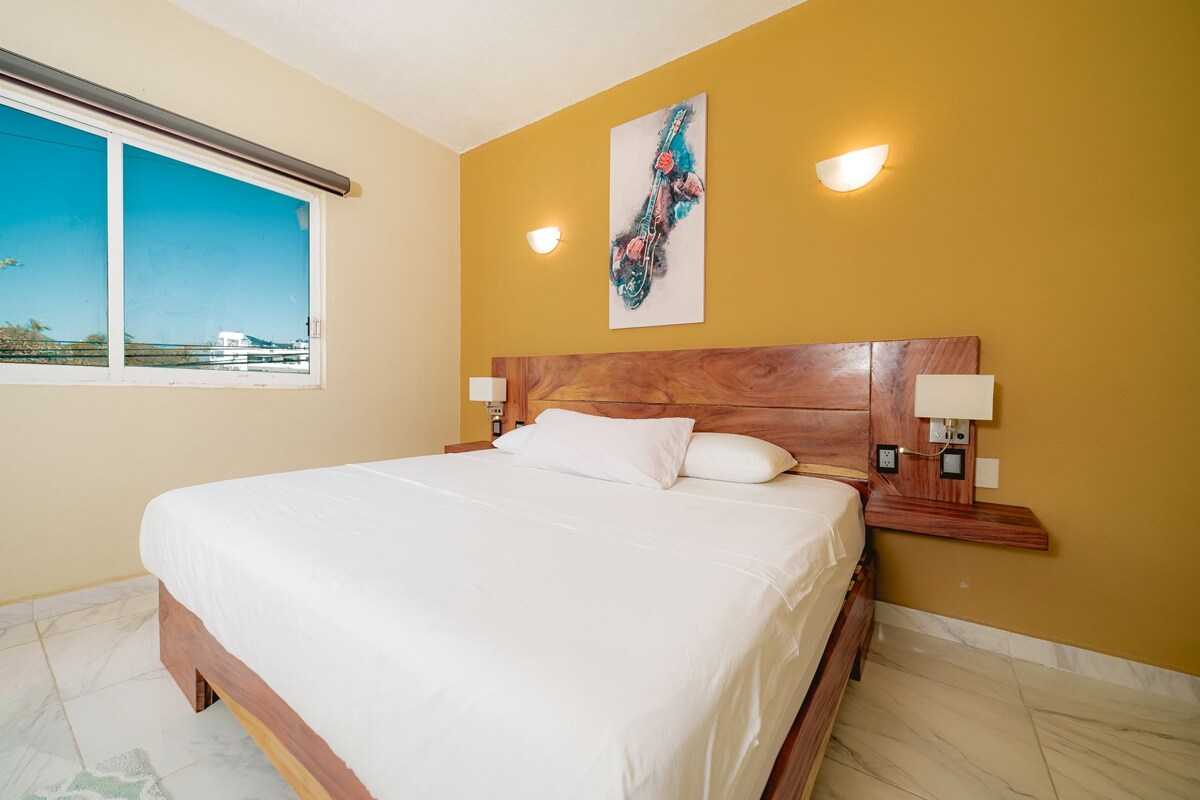 A hotel room with a large bed, a window with a view, a wooden headboard, and two wall-mounted lamps.