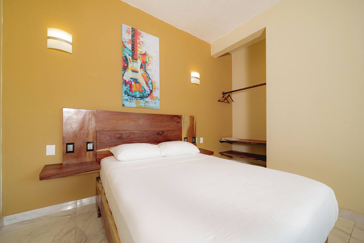 A hotel room with a large bed, wooden headboard, two wall-mounted lights, a painting, and a wooden bench.