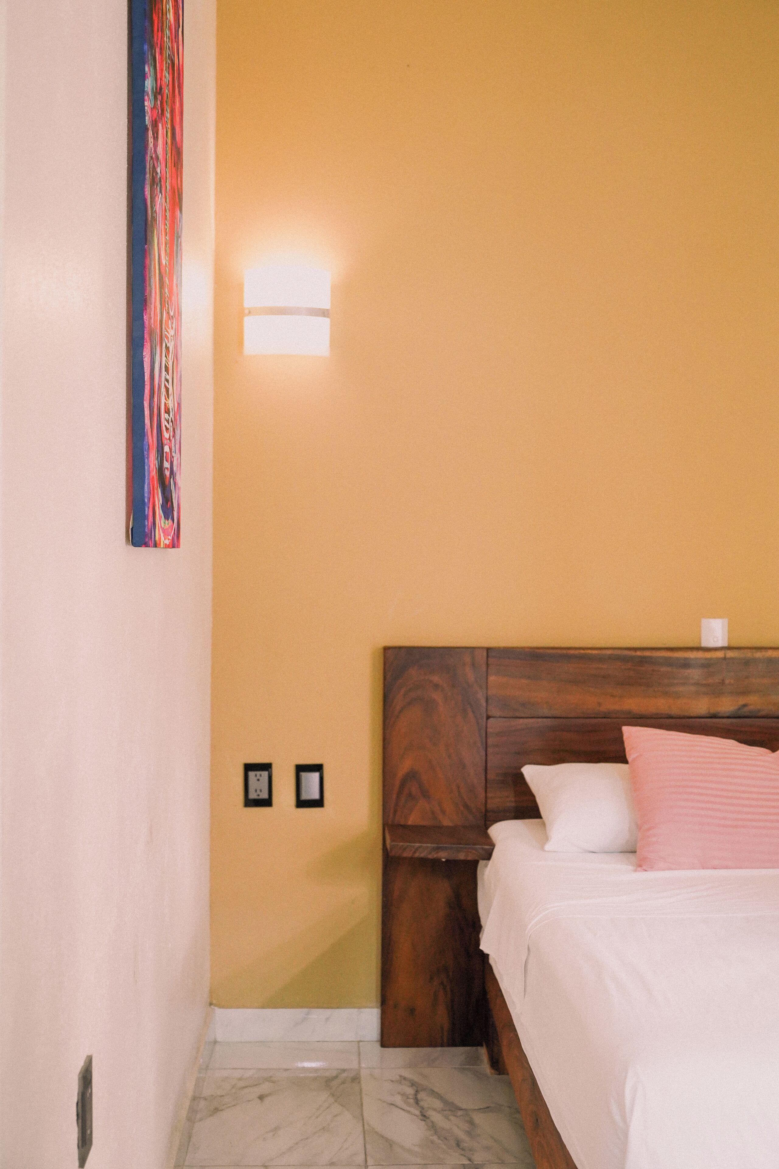 Un dormitorio con cabecera de madera, cama con ropa de cama blanca y rosa, una lámpara de pared y una obra de arte colorida.