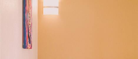 Un dormitorio con cabecera de madera, cama con ropa de cama blanca y rosa, una lámpara de pared y una obra de arte colorida.