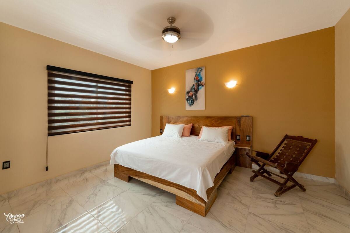 A bedroom with a bed, a wooden headboard, a chair, a ceiling fan, and a painting on the wall.