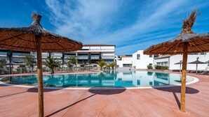 Une piscine agrémentée de parasols en chaume, une terrasse carrelée et, en arrière-plan, des bâtiments modernes.