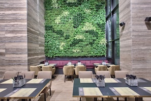 A restaurant with a green vertical garden wall, wooden tables, and beige chairs.