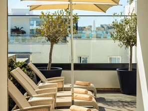 A rooftop terrace with wicker lounge chairs, a large umbrella, and potted plants.