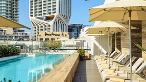 A rooftop pool area with lounge chairs and umbrellas, a modern building with a unique facade, and a clear blue sky.