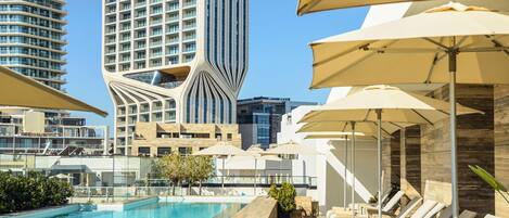 A rooftop pool area with lounge chairs and umbrellas, a modern building with a unique facade, and a clear blue sky.
