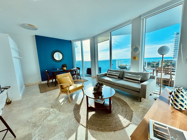 A spacious living room with a large sofa, a coffee table, and a dining area. The room has large windows offering a view of the ocean. - Condo on Miami Beach with stunning views from oversized private terrace. (Miami)