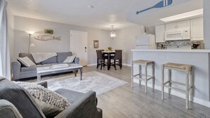 Un salon moderne avec un canapé gris, une table basse et un coin repas comprenant une table et des chaises. On y trouve également une cuisine avec des meubles blancs et un four à micro-ondes.