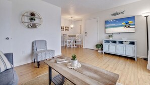 Een woonkamer met een houten salontafel, een grijze fauteuil, een witte eethoek en een televisie aan de muur.