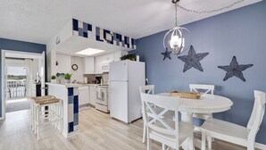 A modern kitchen with white cabinetry, a built-in microwave, and a white refrigerator. There is a dining area with a white table and chairs, and a blue accent wall with star decorations. - Caddy's #5 – Waterfront Balcony, Walk to Beach, Pet-Friendly & Next to Dining (Indian Shores)