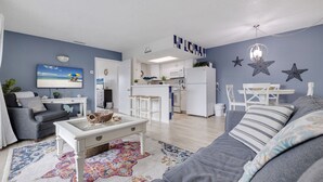 A modern living room with a sofa, coffee table, and a TV. There is a kitchen area with white cabinets and a microwave. - Caddy's #5 – Waterfront Balcony, Walk to Beach, Pet-Friendly & Next to Dining (Indian Shores)