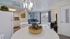 A modern living room with a dining table, white chairs, a chandelier, and a "Gather" sign on the wall.