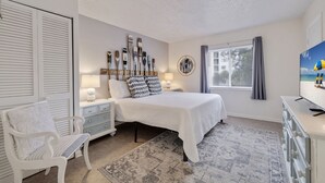 A bedroom with a bed, bedside tables, a chair, a TV, and a window with curtains. - Caddy's #8 – Walk to Beach, Pet-Friendly, Full Kitchen & Steps to Dining (Indian Shores)