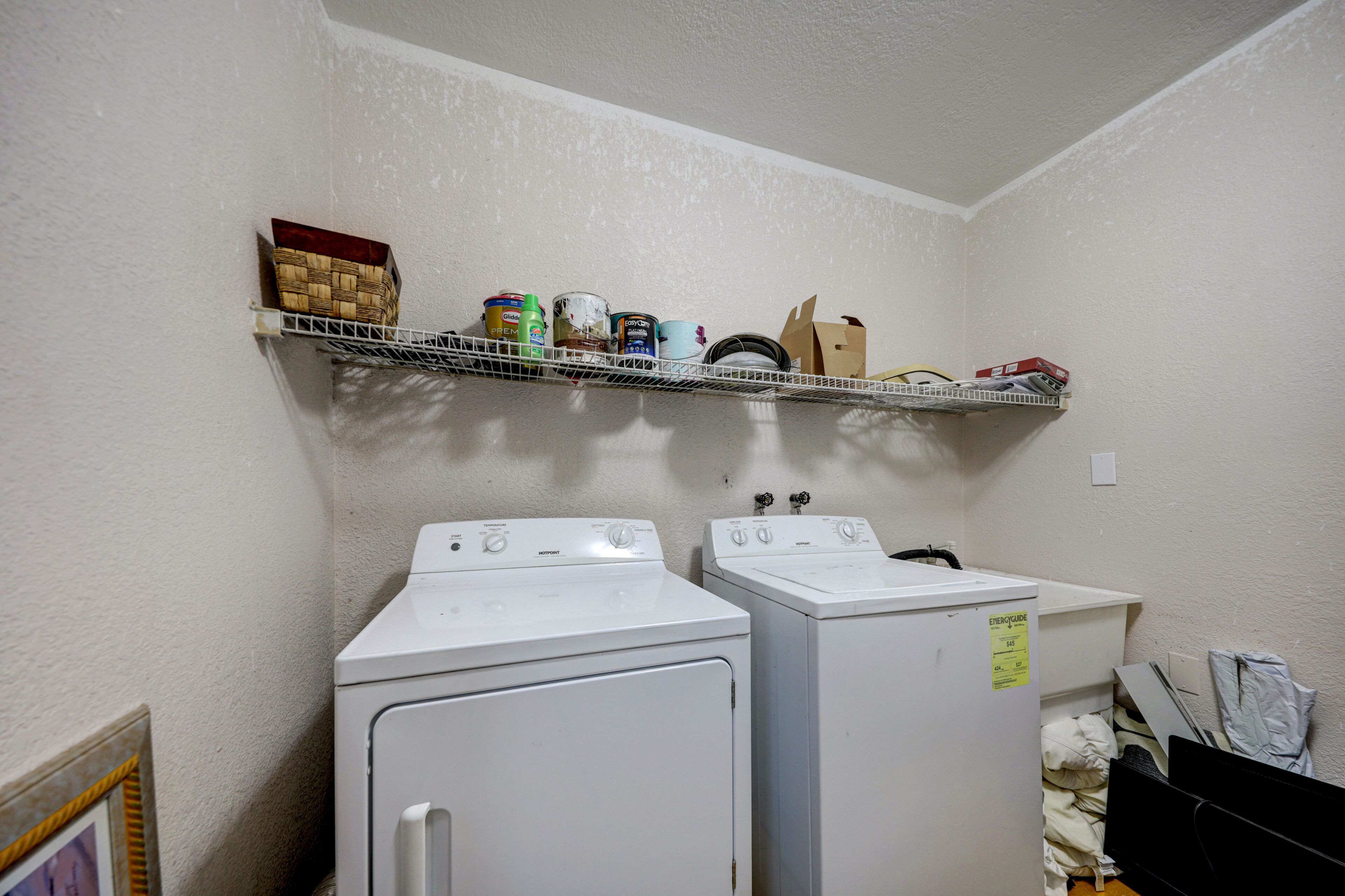 A laundry room with two white front-loading washers and a metal shelf above holding various items.