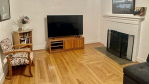 Een woonkamer met een flatscreen televisie op een houten standaard, een open haard, een boekenkast en een fauteuil met bloemenprint.