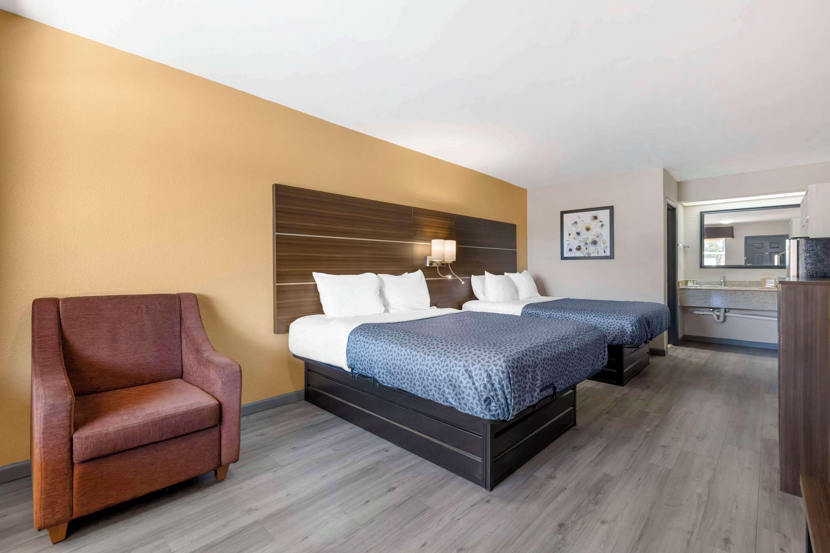 A hotel room with two beds, a sofa, a wooden headboard, a wall-mounted lamp, and a small bathroom with a mirror and sink.