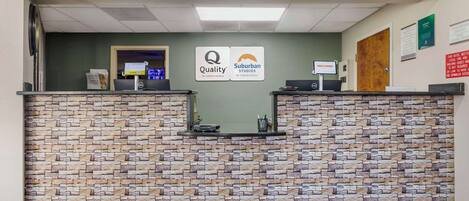 A reception desk with a stone-patterned wall, a computer, and a sign indicating 'Quality by Choice Hotels' and 'Suburban Studios by Choice Hotels'.