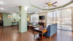 A spacious living area with a wooden floor, a ceiling fan, a flat-screen TV, and a large window with sheer curtains. - Suburban Studios Orangeburg (Orangeburg)