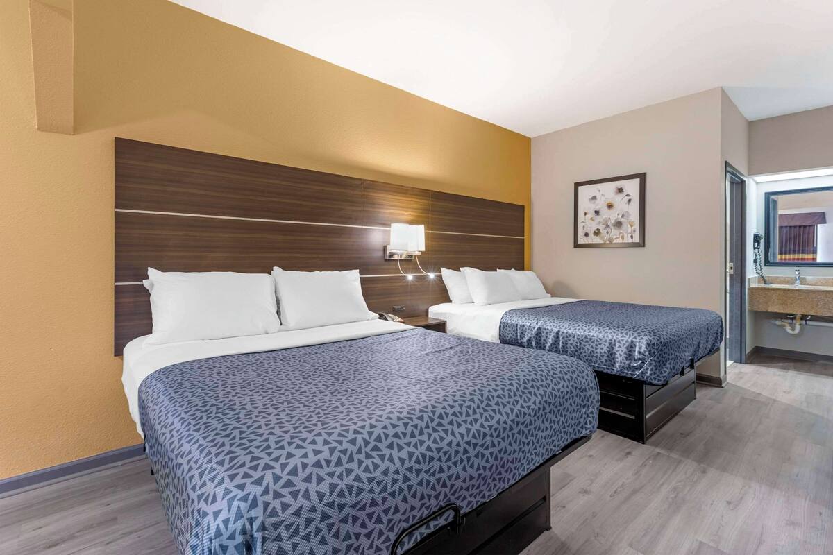 A hotel room with two beds, a wooden headboard, a nightstand, a wall-mounted lamp, and a framed picture.