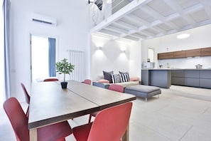 Un salon moderne comprenant une table à manger, des chaises rouges, un canapé gris et un coin cuisine doté d’un plan de travail gris et de meubles de rangement en bois.