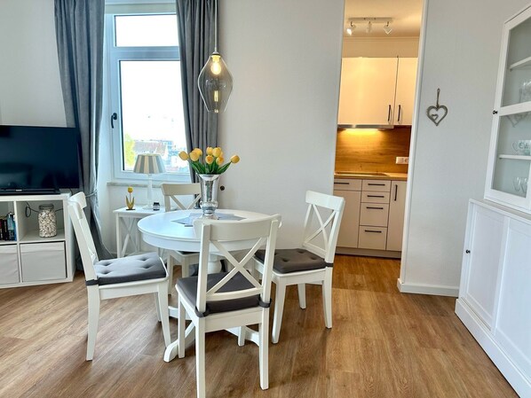 Ein modernes Wohnzimmer mit einem runden Esstisch, weißen Stühlen, einem Fernseher und einer Vase mit gelben Blumen.