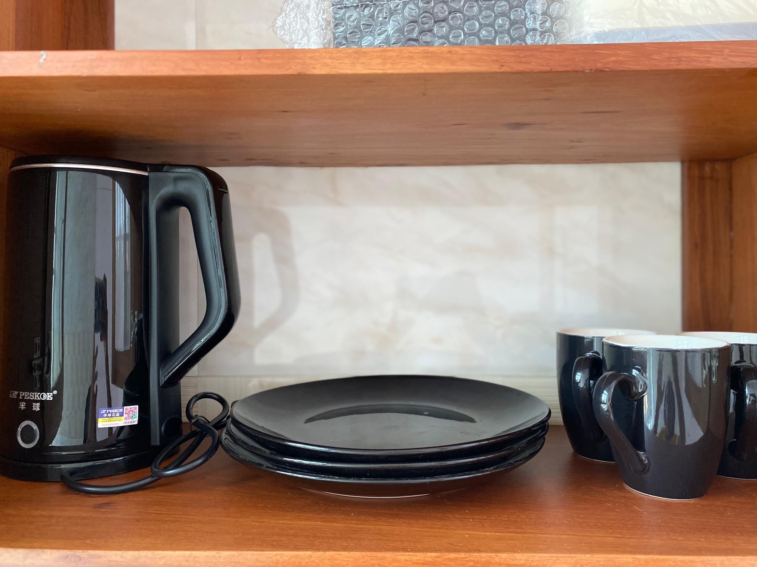 Una tetera eléctrica negra, una pila de platos negros y tazas negras sobre un estante de madera.