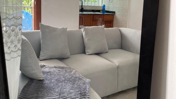 A modern living room with a light grey sofa, a patterned throw blanket, and a kitchen area with wooden cabinets and a window. - LuckySmallie 1-Bed Apartment in Goba,Dar es Salaam (Dar es Salaam)