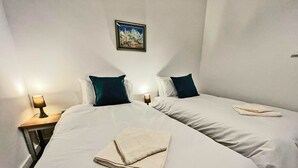 Habitación de hotel con dos camas, ropa de cama blanca, almohadas oscuras, un cuadro en la pared y una toalla doblada sobre la cama.