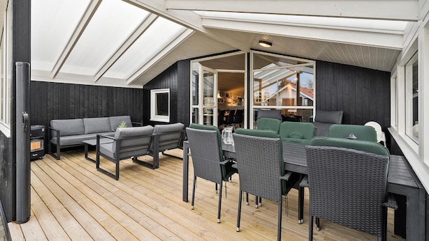Ein überdachter Außenbereich mit Holzterrasse, grau-grünen Möbeln und Blick ins Haus durch Glastüren.