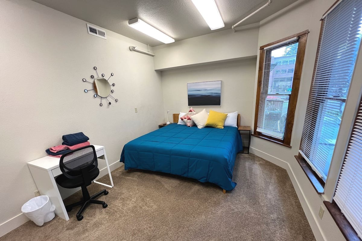 Un dormitorio con cama, escritorio, silla, cesto de basura y ventana con persianas.