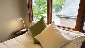 Ein Hotelzimmer mit Bett, Kissen, Nachttisch, Lampe und einem Fenster mit Blick auf Bäume und ein Gebäude.