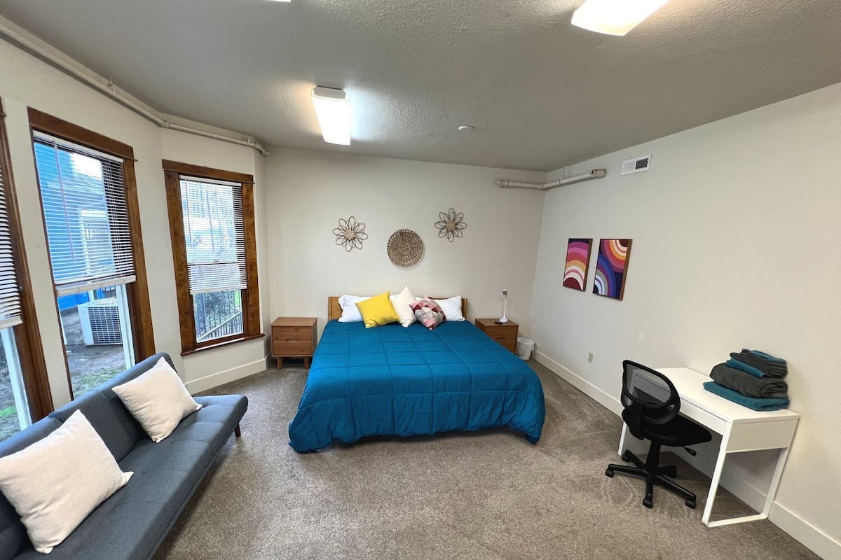 Un dormitorio con cama, escritorio, sillón, sofá y dos ventanas con persianas.